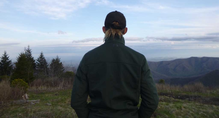 A person facing away from the camera looks out over the vast blue ridge mountains. 